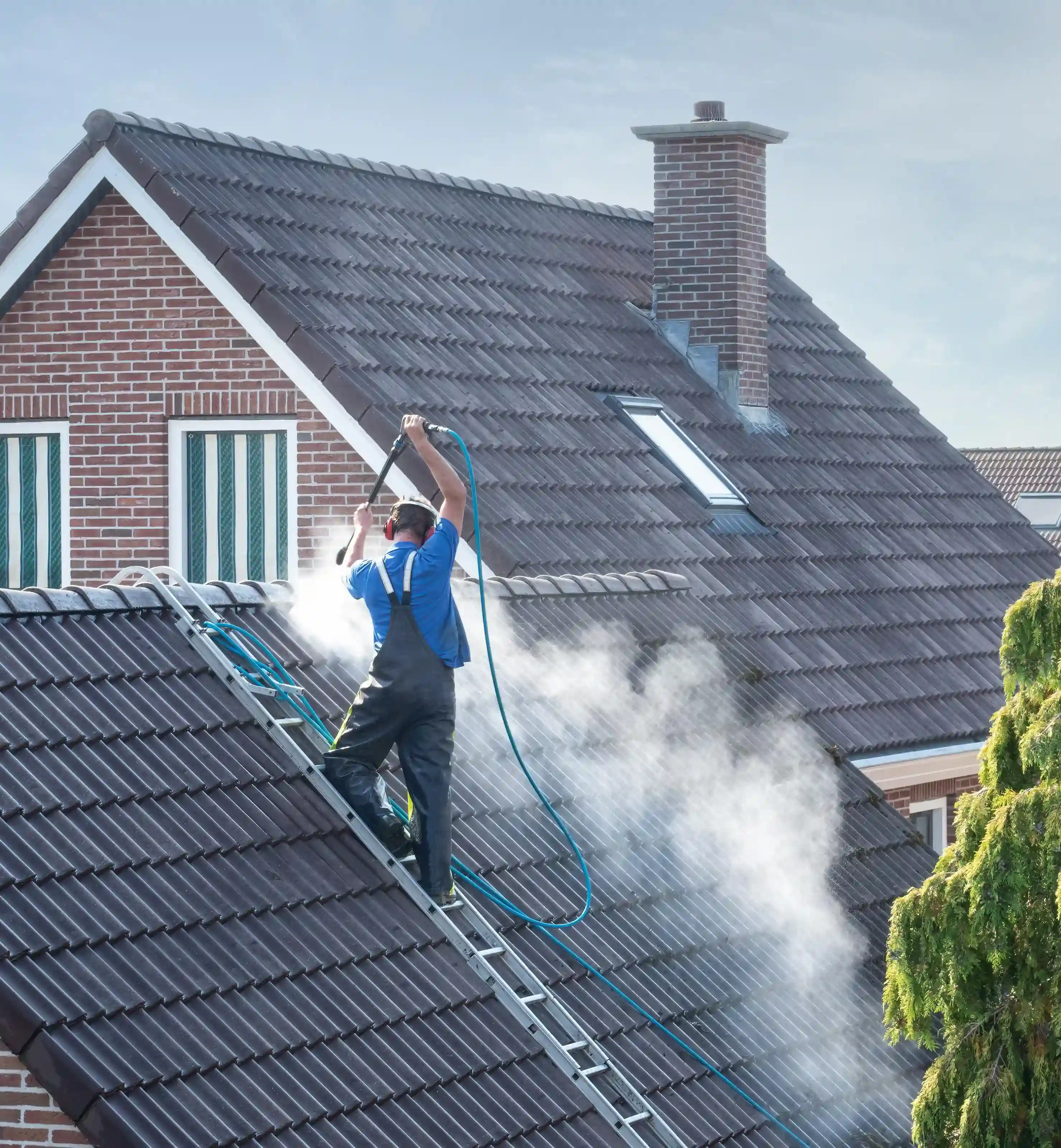 Professionelle Dachreinigung in Aktion: Ein Arbeiter auf einer Leiter entfernt gründlich Schmutz und Ablagerungen von einem Ziegeldach mittels Hochdruckreiniger und Wasserdampf, wobei eine große Sprühnebelwolke entsteht. Im Hintergrund ist ein Backsteinhaus unter blauem Himmel zu sehen.