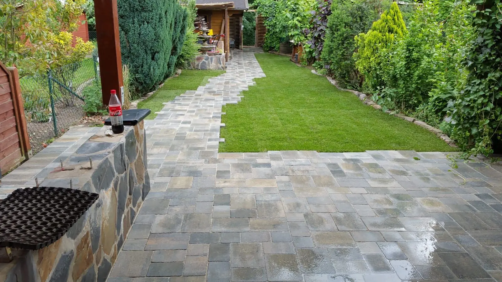 Terrassenbau NRW Nachher: Moderne Terrasse mit hochwertigen grauen Pflastersteinen, Steinmauer und frischem Rollrasen, umgesetzt in Nordrhein-Westfalen.